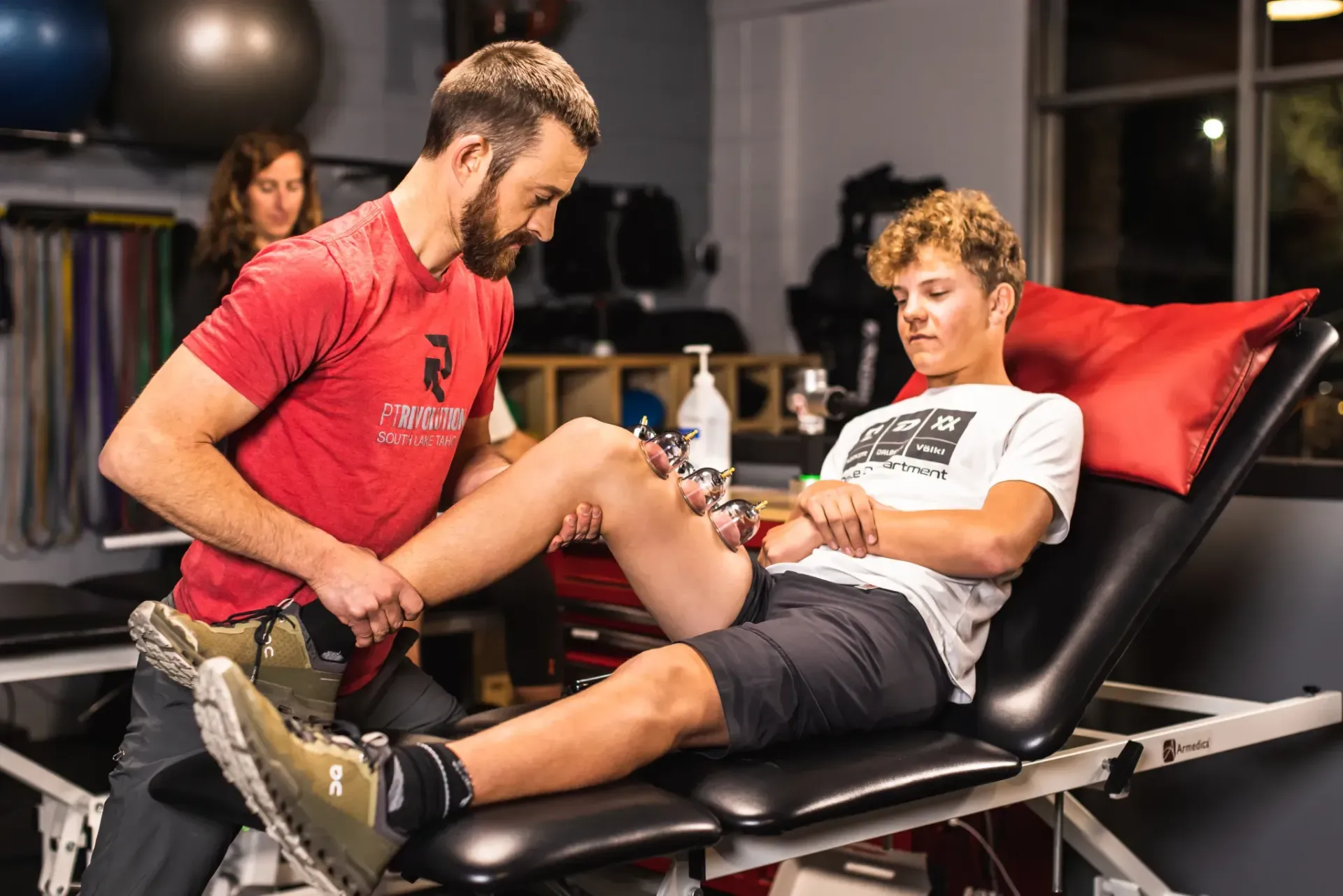 Trainer stretching athlete’s leg at PT Revolution on a treatment table in a gym, with a woman in the background