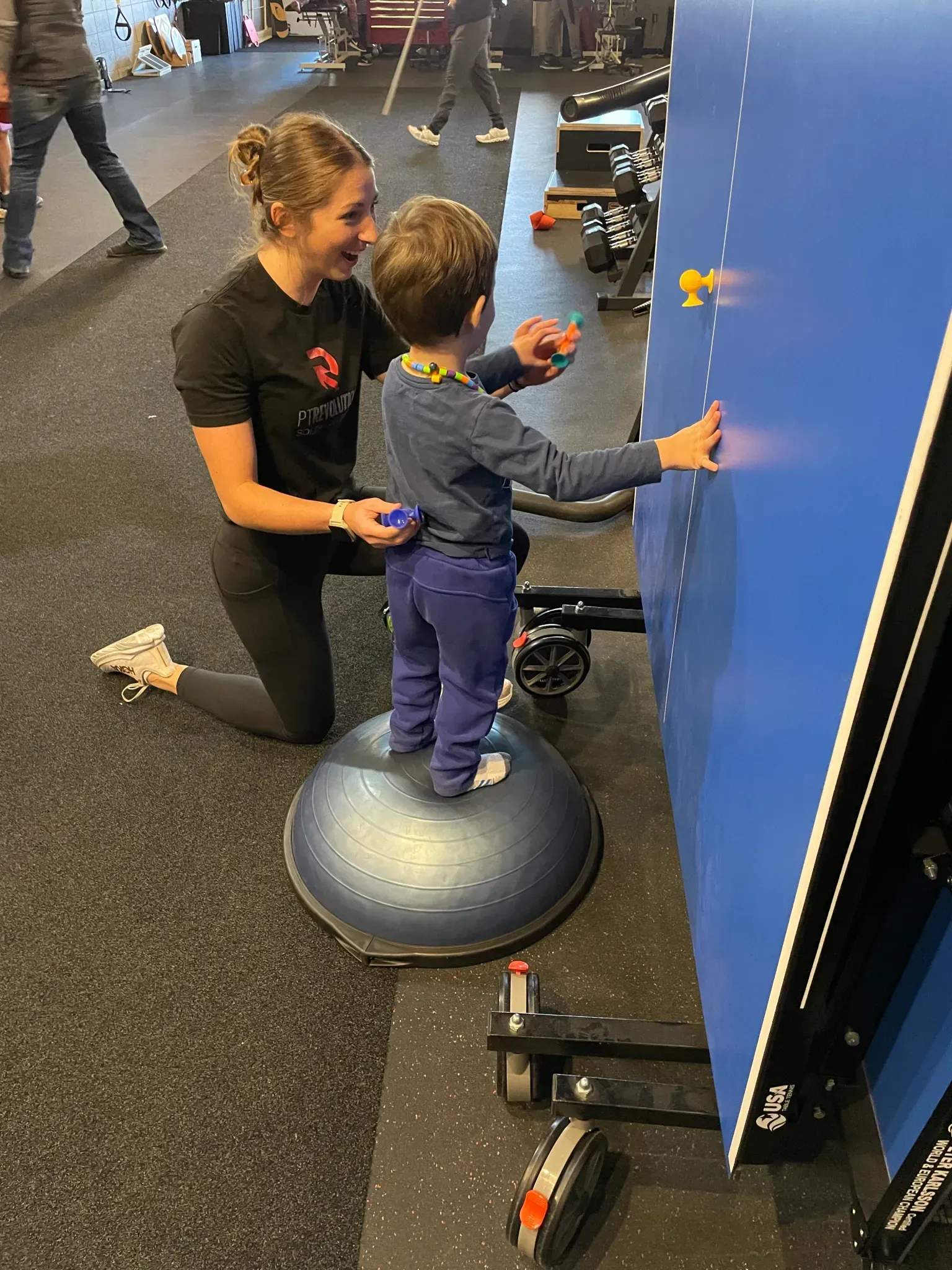 Child and instructor use a blue interactive wall game in a gym, standing on a balance dome at PT Revolution.