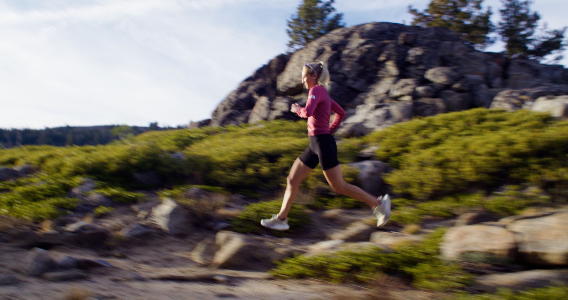 Runner in a pink shirt jogging along a rocky trail in a sunny, wooded landscape