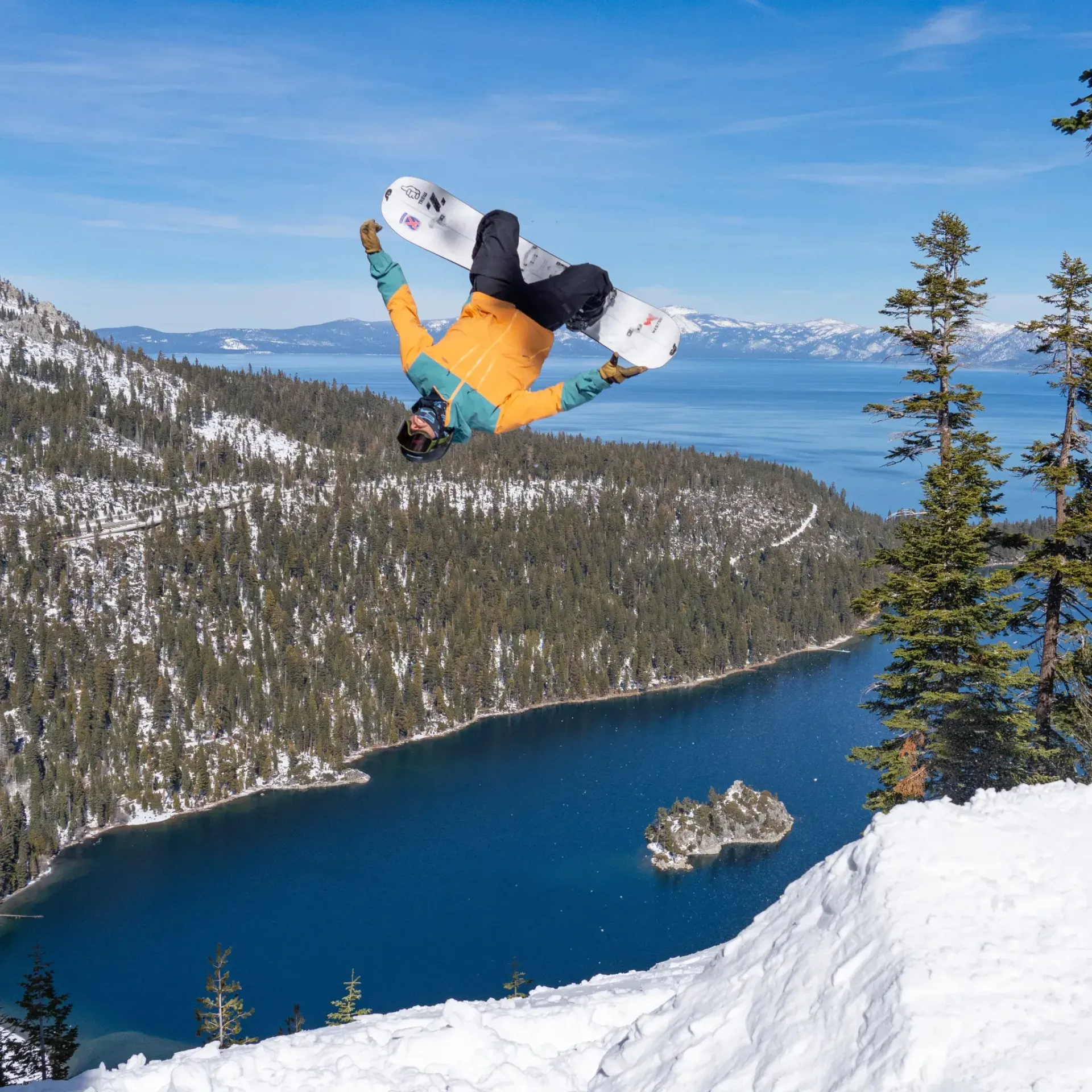 Snowboarder airborne above a snowy lake and pine-covered mountains under a blue sky