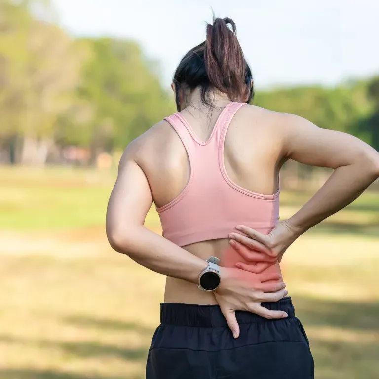 Person in pink tank top holding lower back while standing outdoors in a park