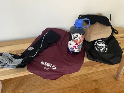 Water bottle and clothing items on a wooden bench, including a maroon jacket and black bag.