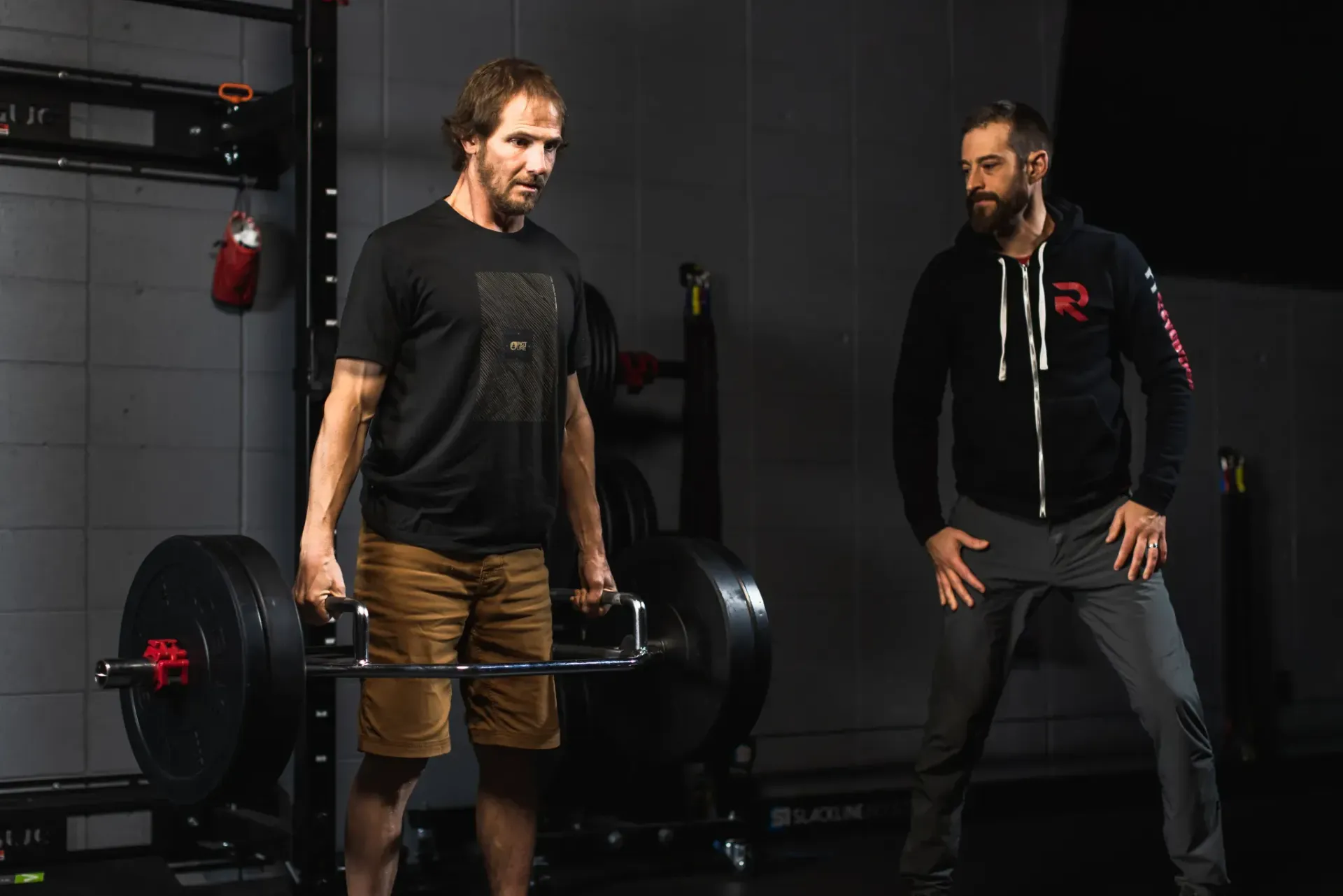 Two men in a gym preparing to lift a heavy barbell, one wearing headphones and lifting gloves.