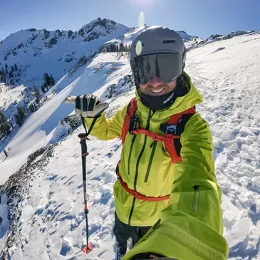 Skier in neon jacket holding poles on a snowy mountain ridge under bright sun
