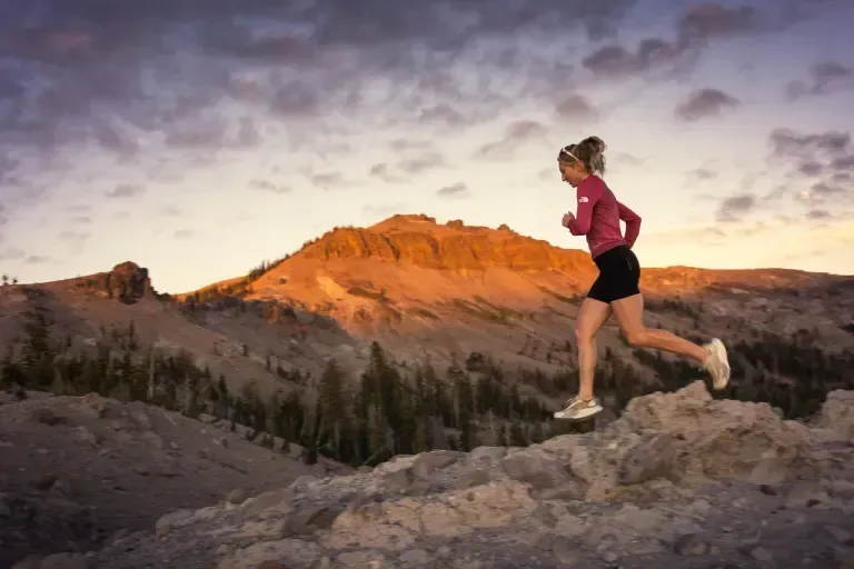 Runner in a pink jacket runs along rocky trail at sunset with orange mountains in the background