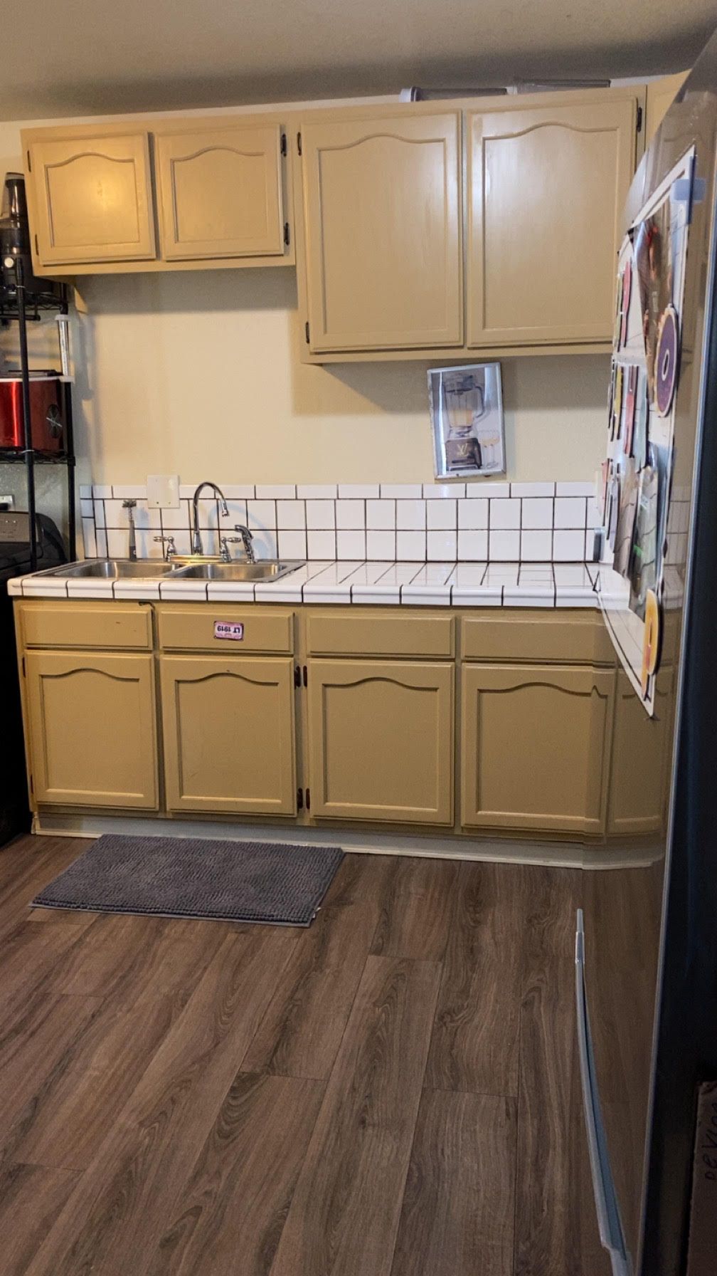 A kitchen with tan cabinets, white tiled backsplash, and a stainless steel sink.  The floor is a wood-like laminate.