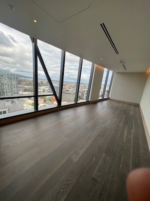 An empty room with dark wood flooring and floor-to-ceiling windows showing a city view.