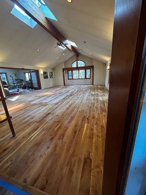 A spacious, sunlit room featuring warm-toned wooden floors, vaulted ceilings with exposed beams, and a large arched window.