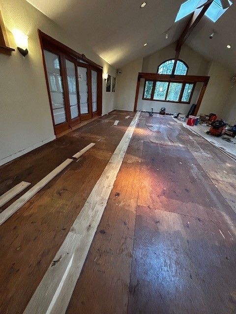 A room under renovation with partially installed hardwood flooring, large windows, and exposed subfloor planks.
