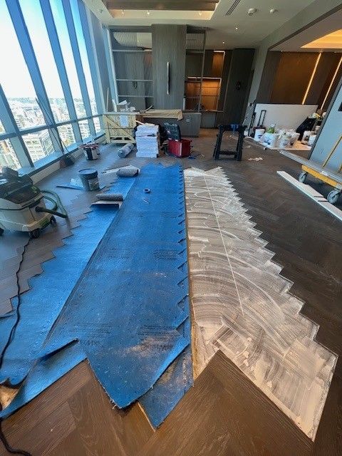 An interior construction site features a room with a herringbone wood floor, blue protective sheeting, and adhesive.