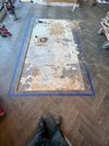 A rectangular section of parquet flooring has been removed, revealing the subfloor, outlined by blue painter's tape.
