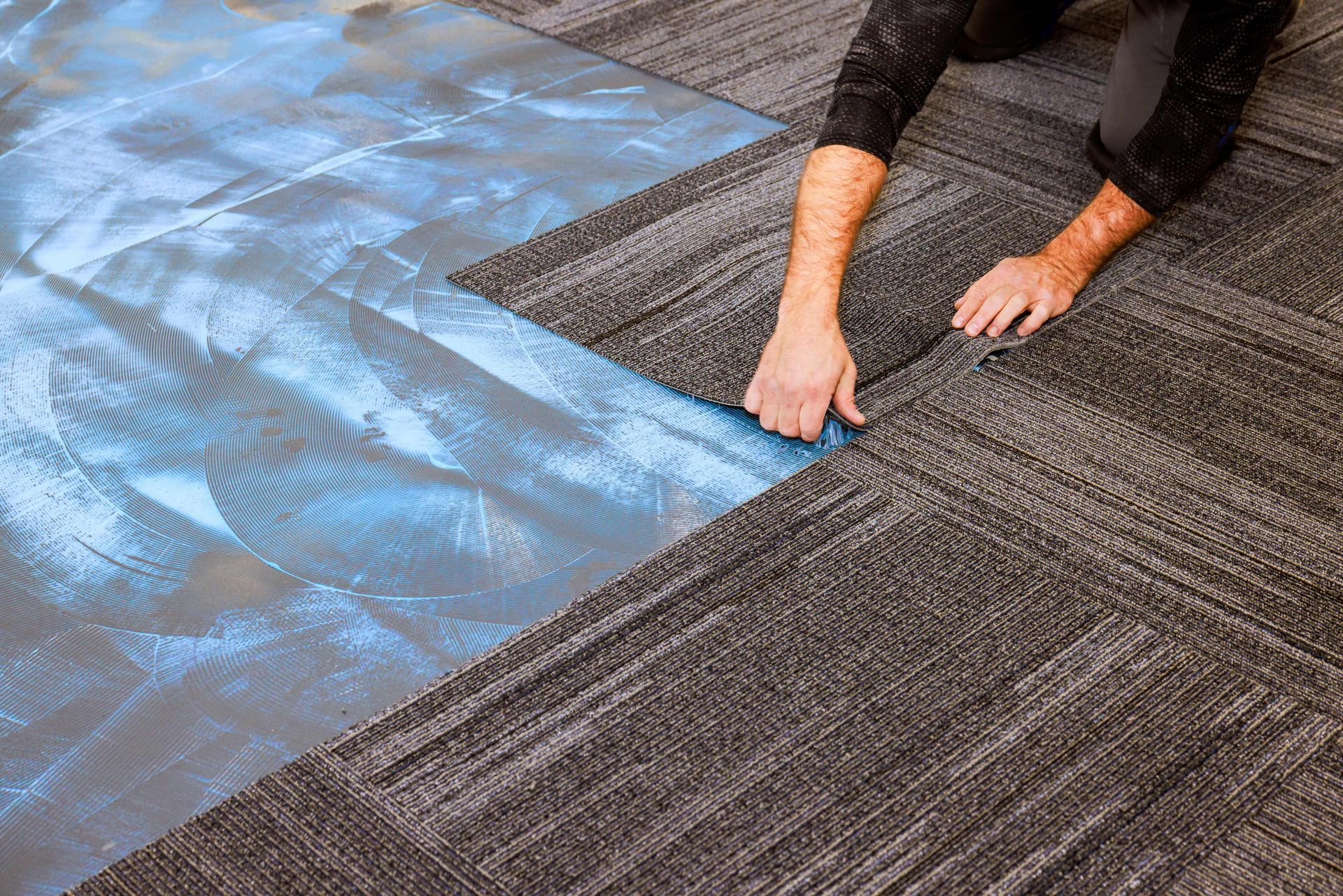 A person installs square carpet tiles onto a floor with a blue adhesive backing. A person installs square carpet tiles onto a floor with a blue adhesive backing.