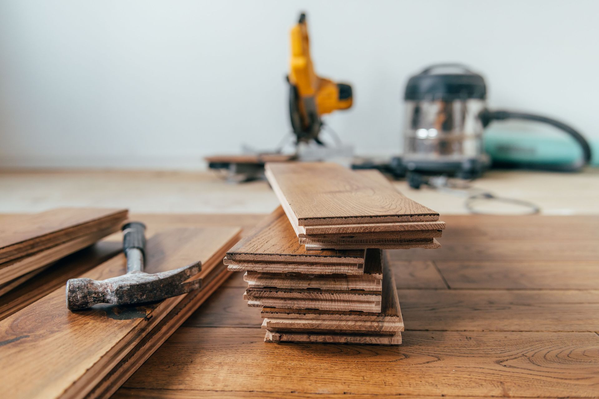 A hammer rests on wood flooring planks next to a stacked pile of flooring, with a power tool and vacuum in the background.