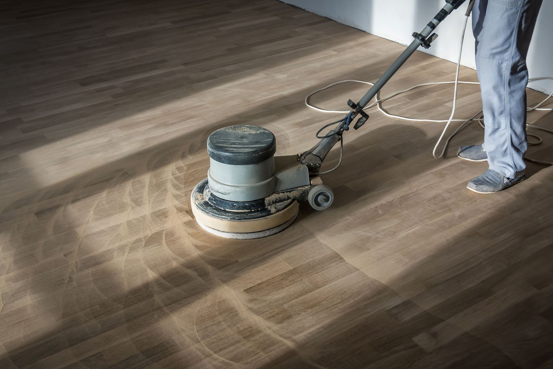 A person uses a floor buffer machine to sand and refinish a light-colored wooden floor.