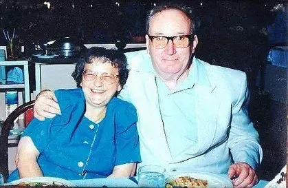 Two people smiling at a table, one in a blue suit with an arm around the other.