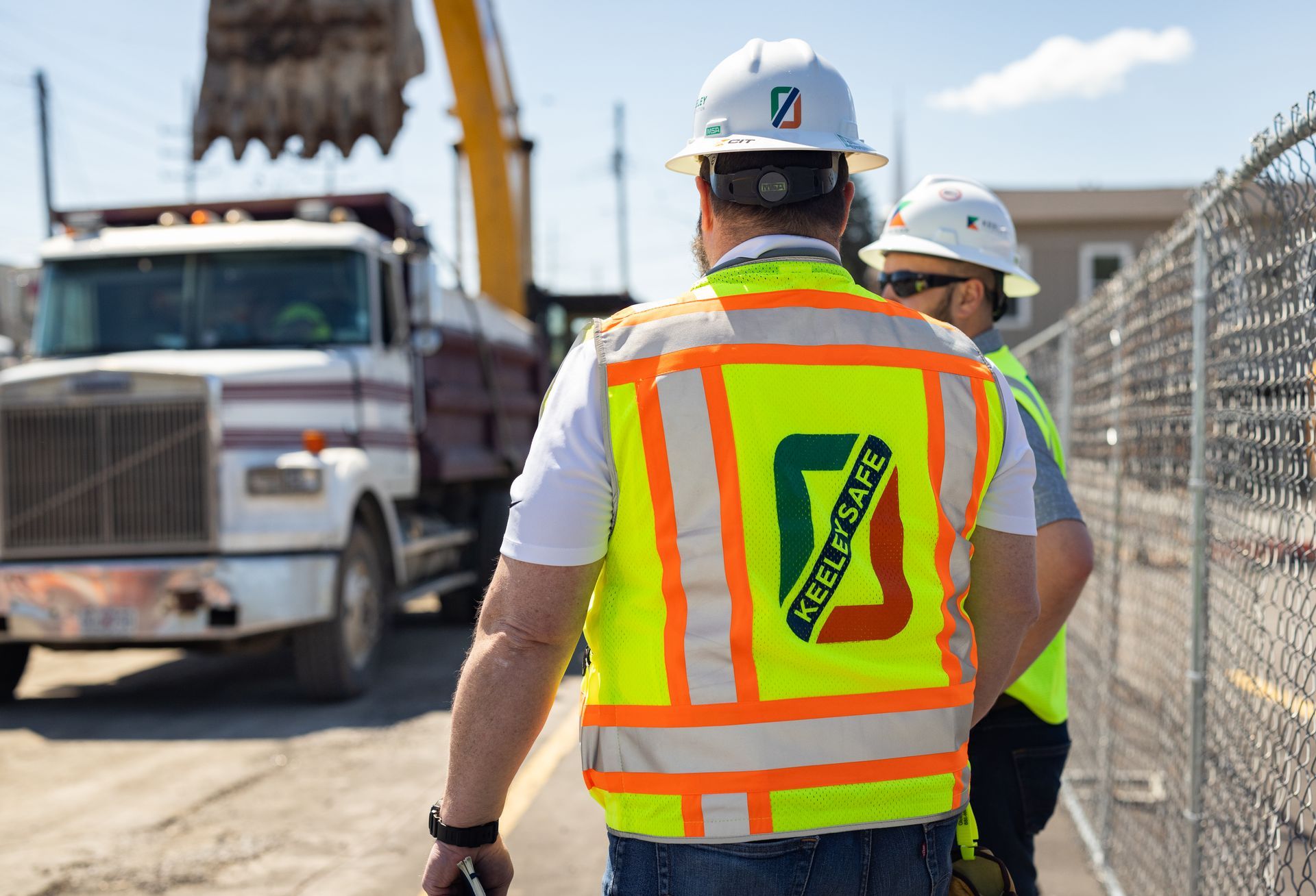 Two construction workers wearing hard hats and safety vests are standing in front of a dump truck.