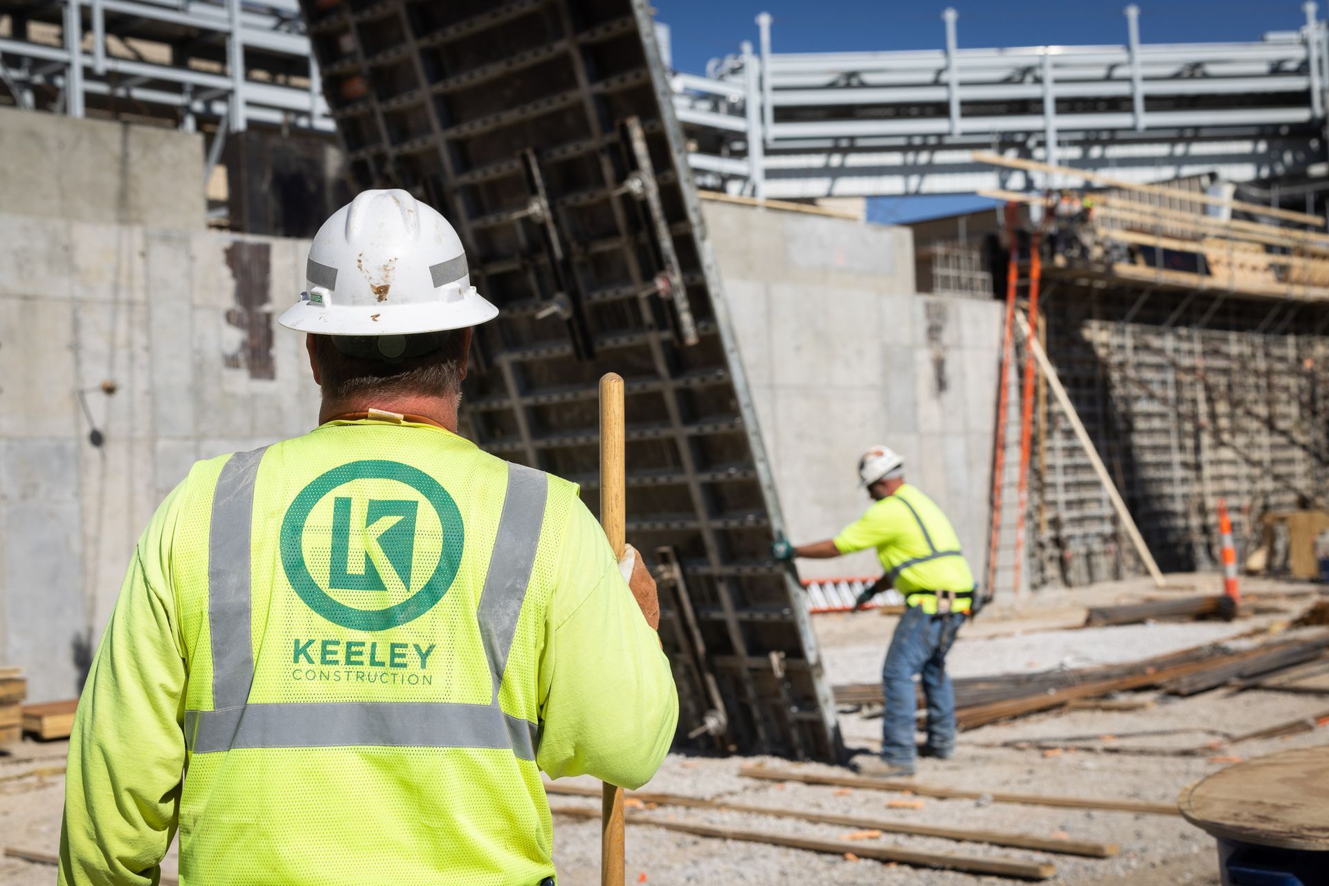 A construction worker wearing a yellow vest with the letter k on it is standing in front of a building under construction.