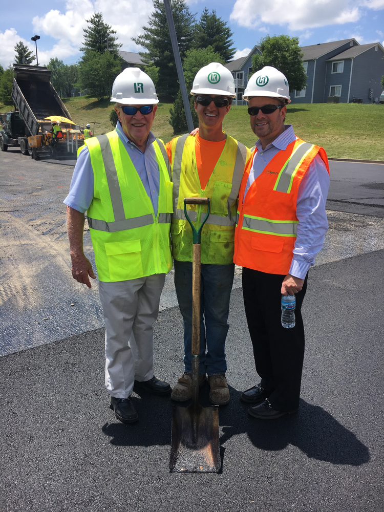 Three men wearing hard hats and safety vests stand next to a shovel