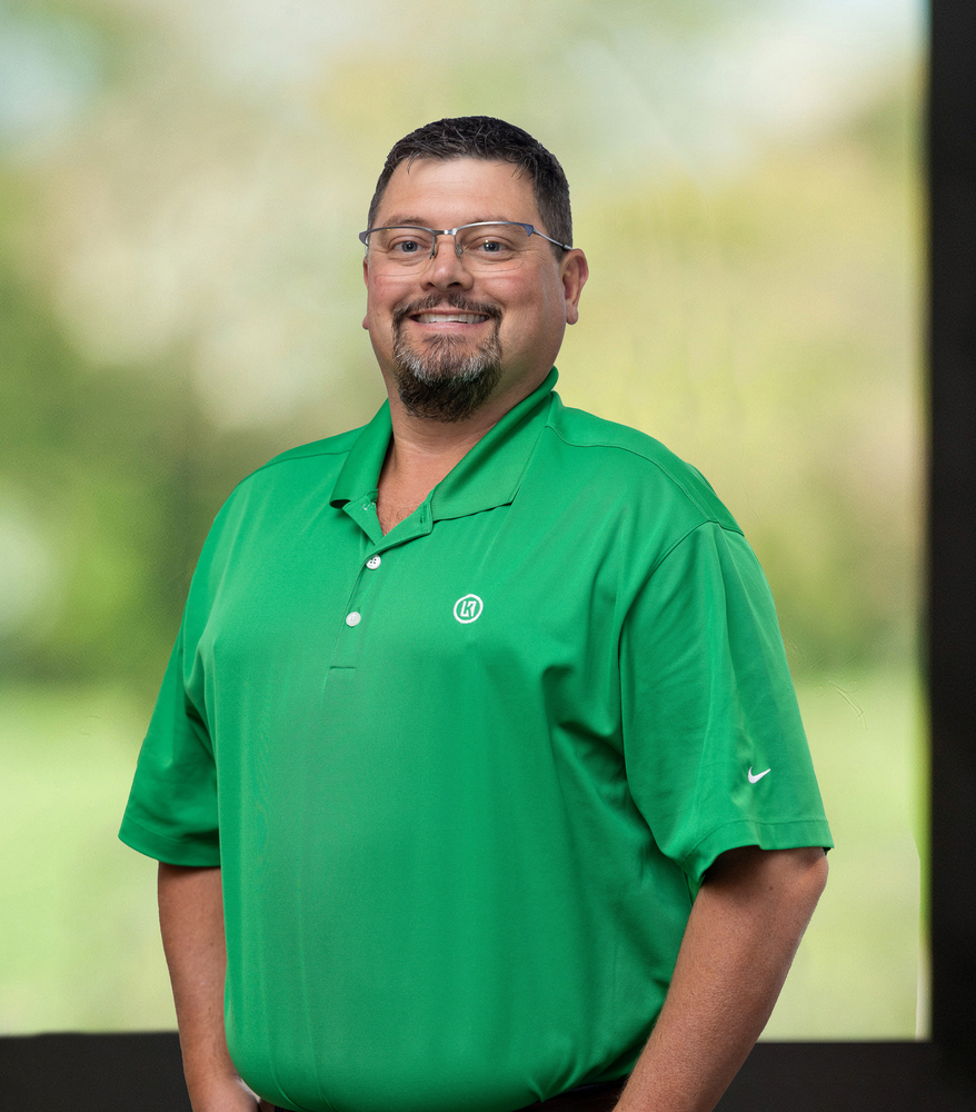 A man wearing glasses and a green shirt is standing in front of a window.