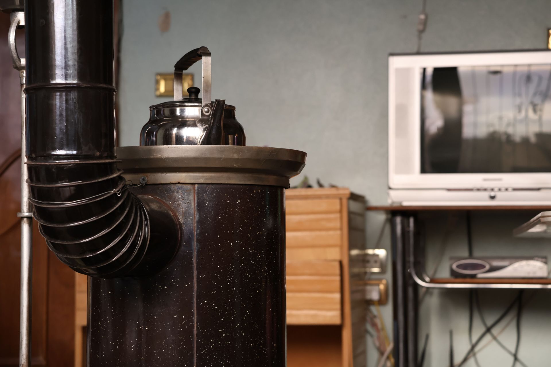 Black wood stove with silver kettle and black vent pipe in a room, with a microwave in the background