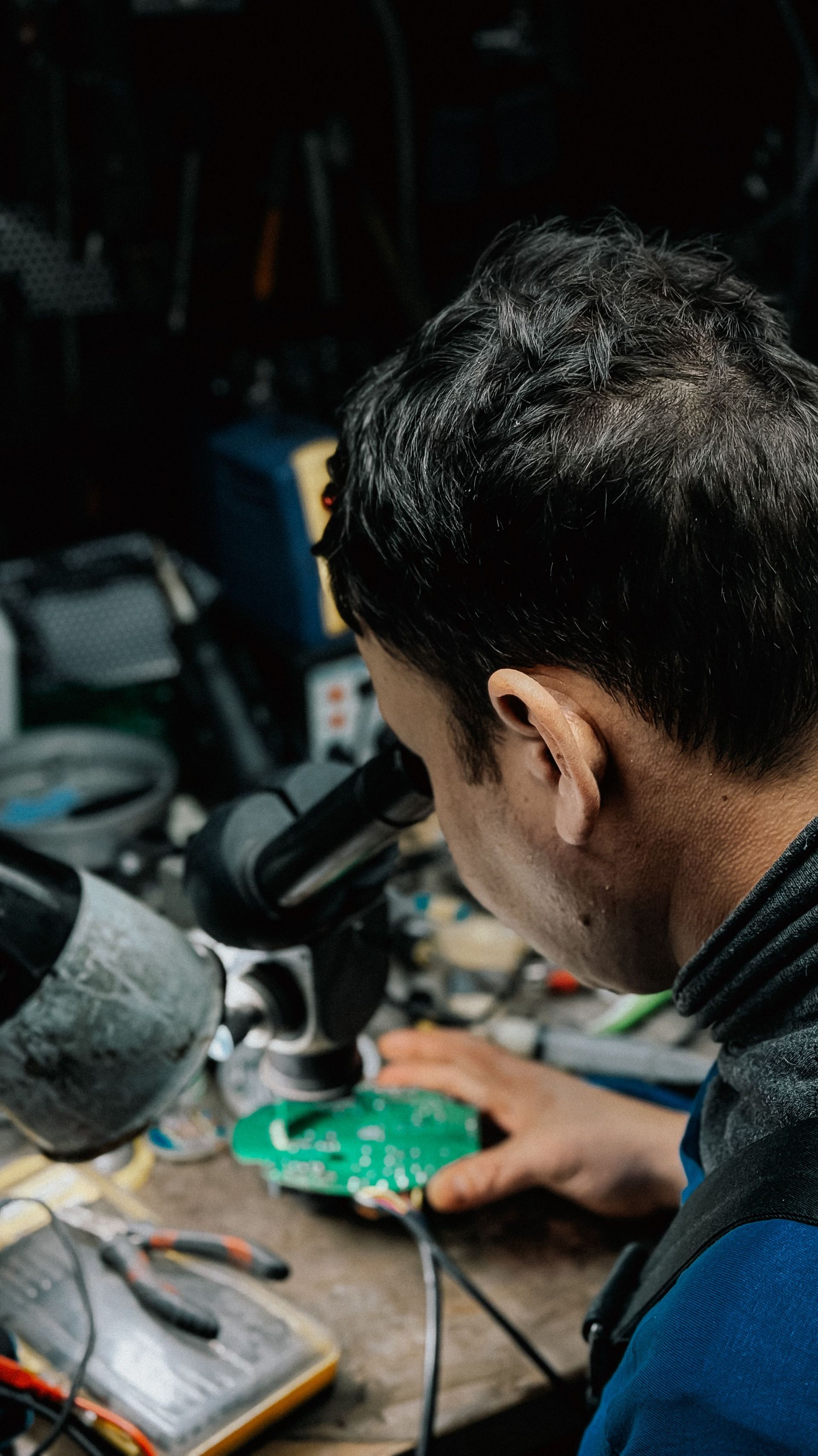 Person working at a cluttered electronics bench, using a magnifier on a green circuit board.