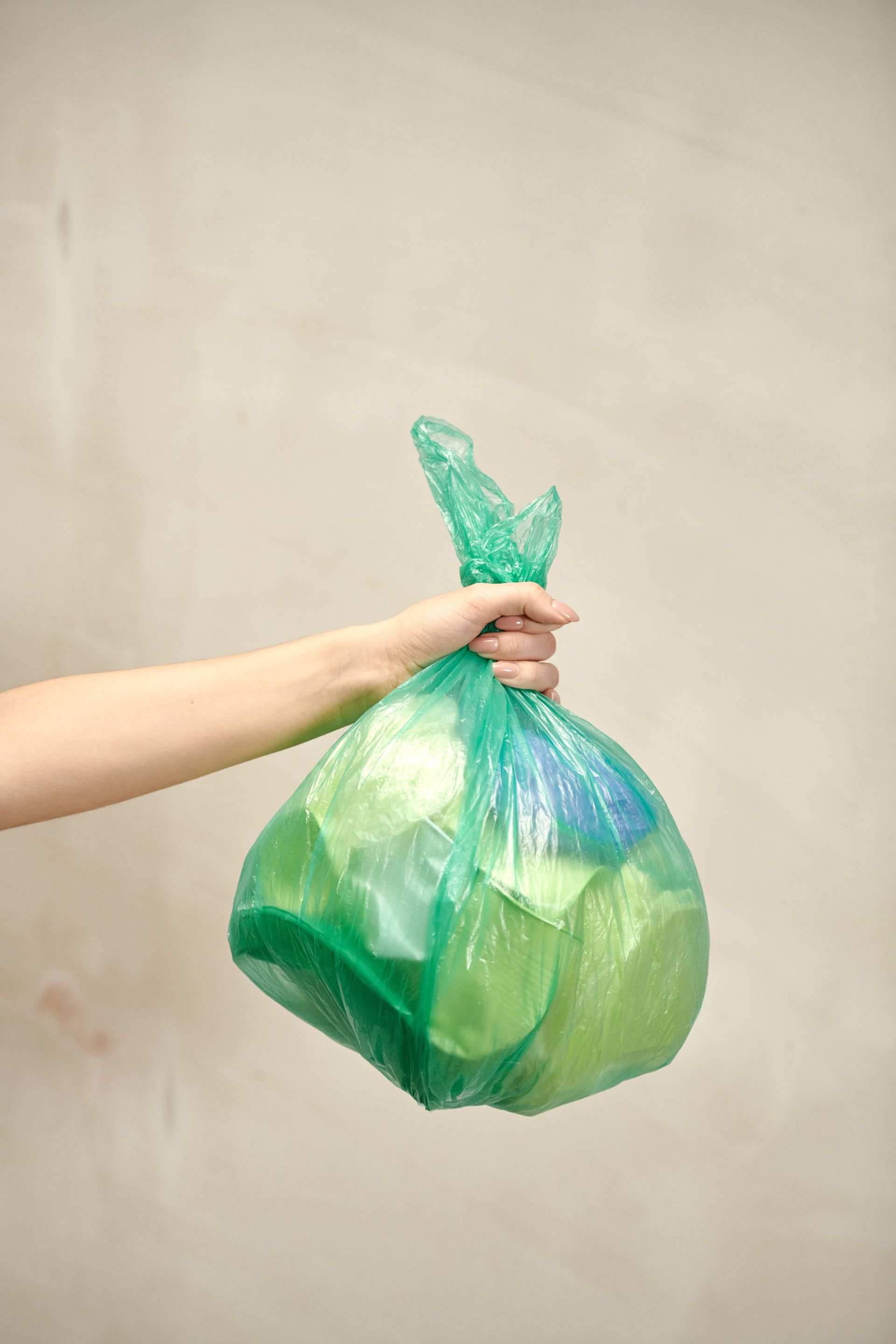 Hand holding a green plastic bag against a plain beige background