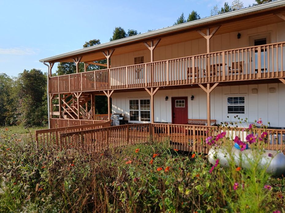 A two-story white building with a wooden wrap-around deck, balconies, and a garden in the foreground under a clear sky.