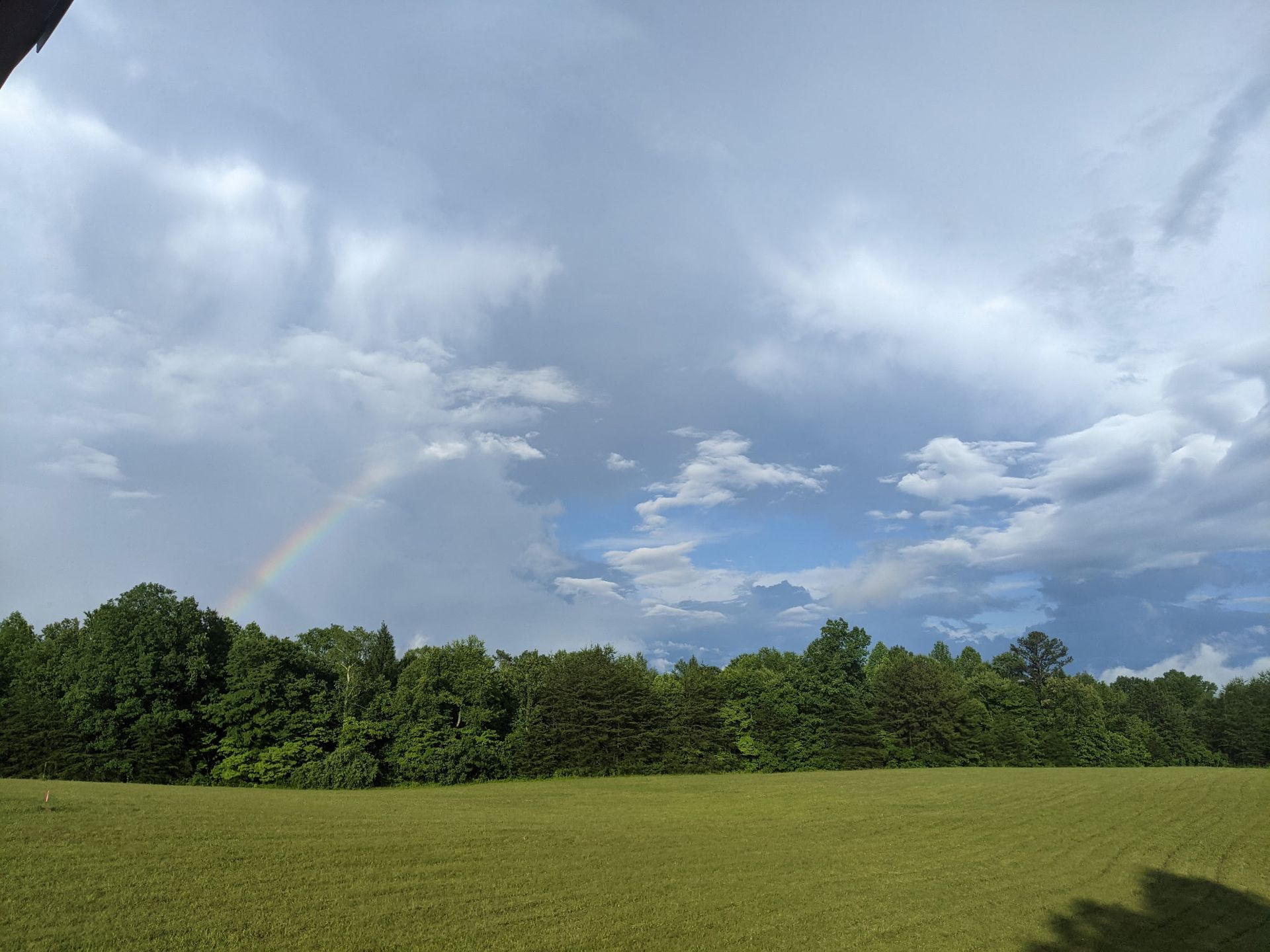 A faint rainbow appears in the sky above a lush green forest and a wide field under a cloudy, bright blue sky.