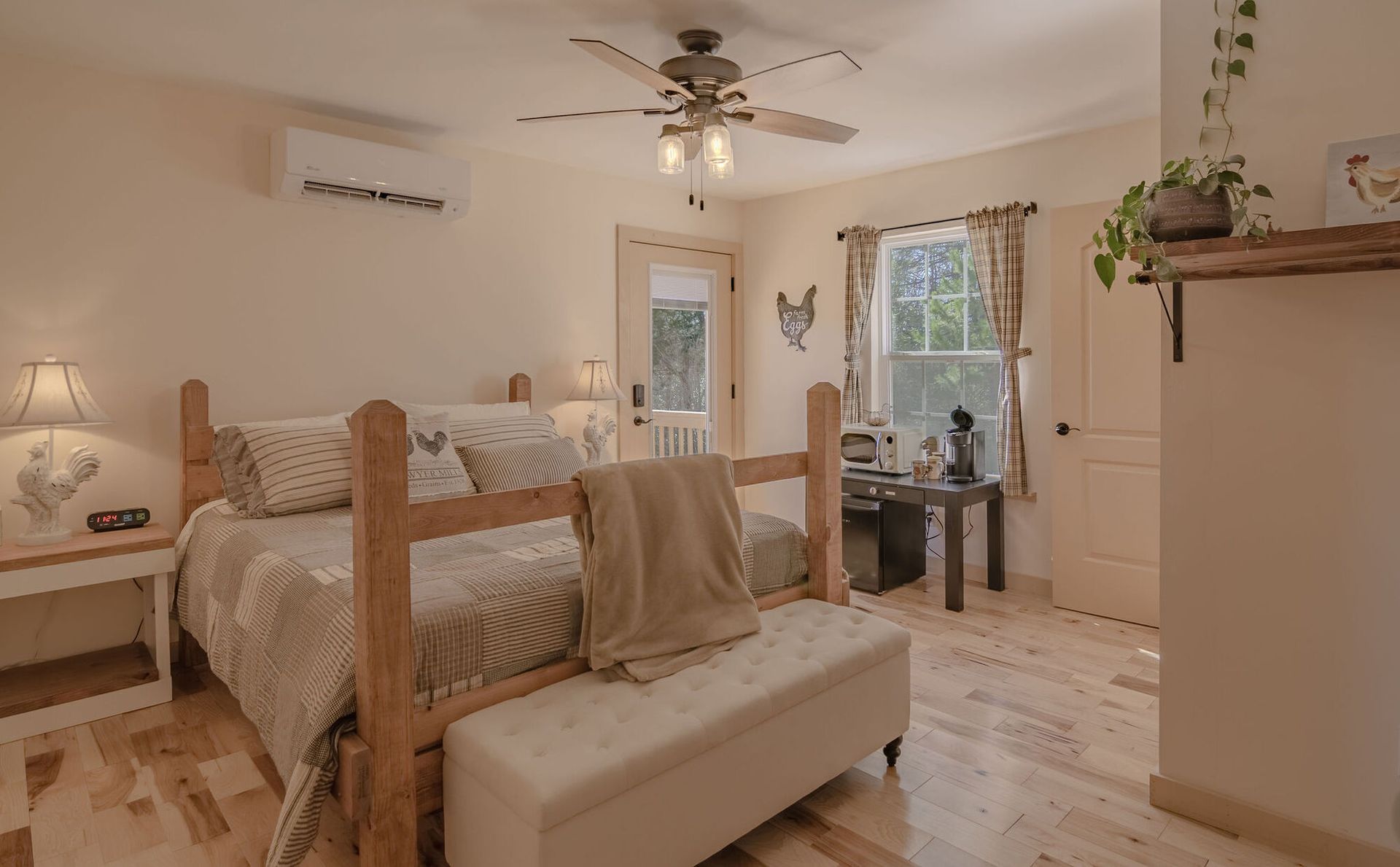 A cozy bedroom features a wooden bed frame, a white bench, a ceiling fan, and a workspace by a window with soft decor.