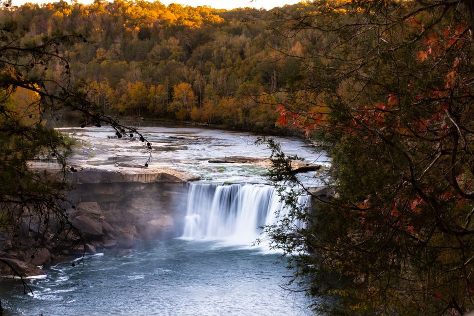 A wide waterfall flows over a rocky ledge into a river, framed by trees with autumn foliage at golden hour.