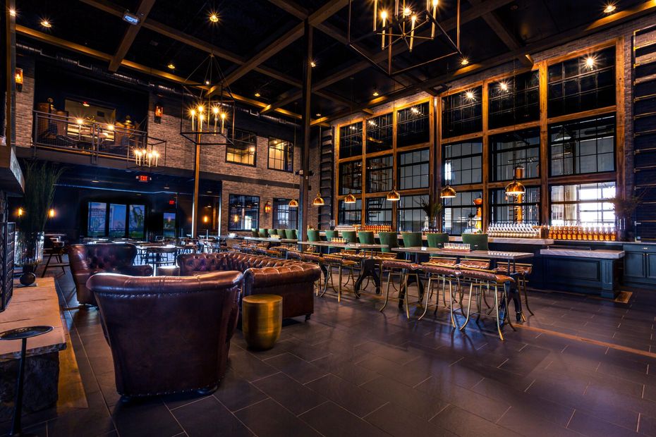 A dimly lit, upscale lounge with dark wood, leather sofas, chandeliers, and high windows overlooking a city skyline.