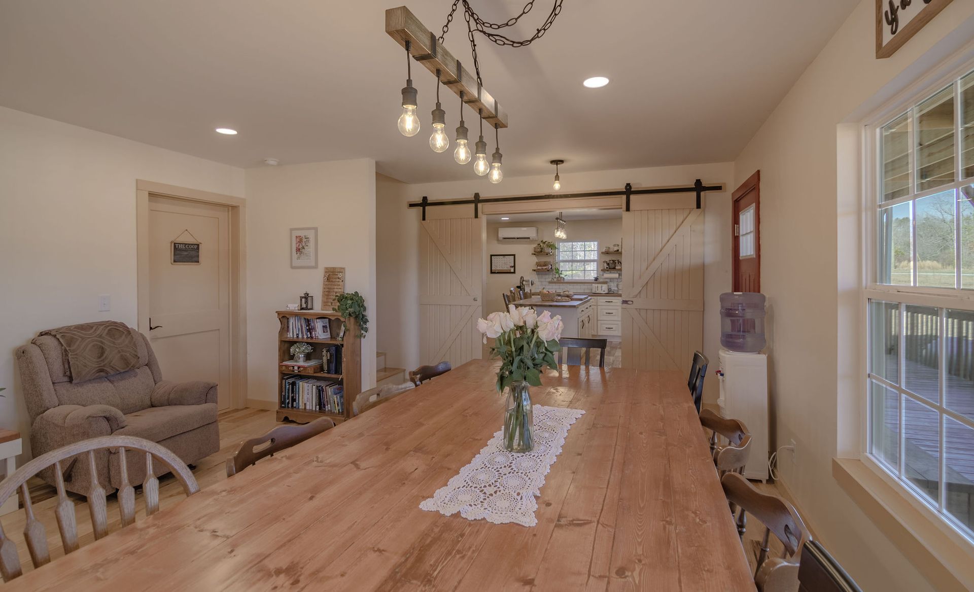 A long wooden dining table sits in a bright room with a rustic pendant light, farmhouse decor, and an open doorway.