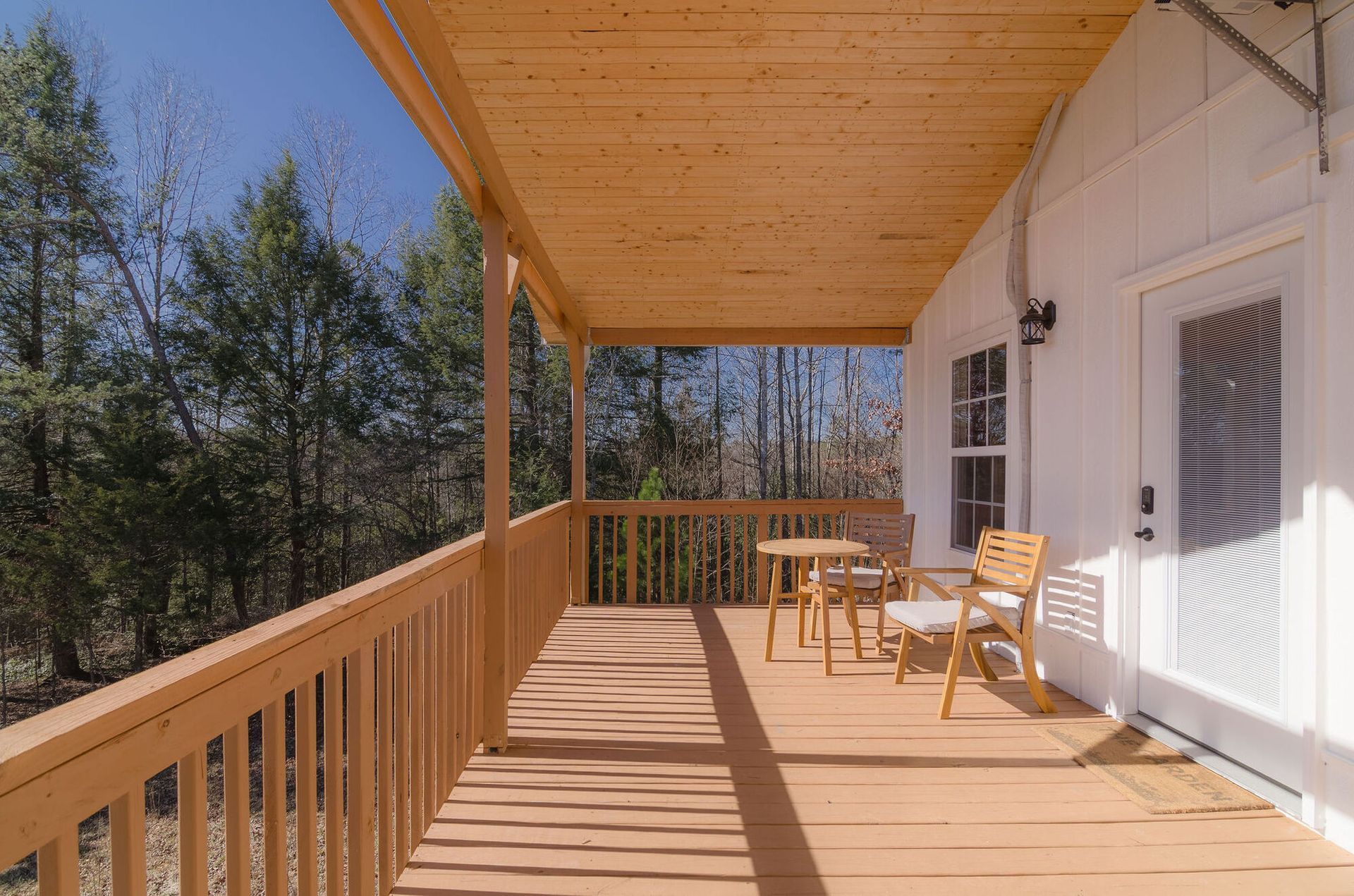A wooden porch with a table and two chairs, overlooking a line of trees on a sunny day.