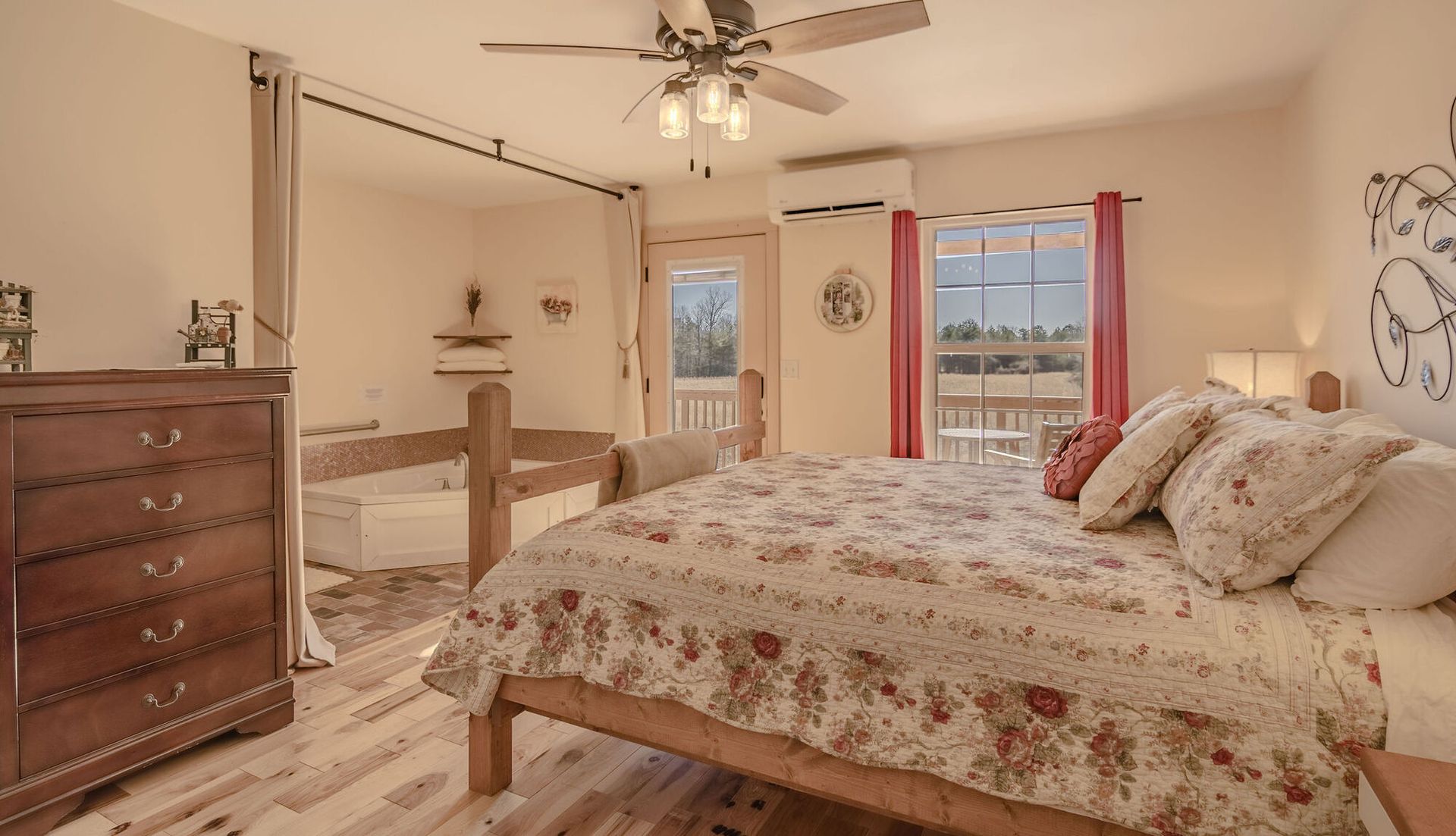 A bedroom with a large bed featuring a floral quilt, a wooden dresser, a ceiling fan, and an attached garden tub area.