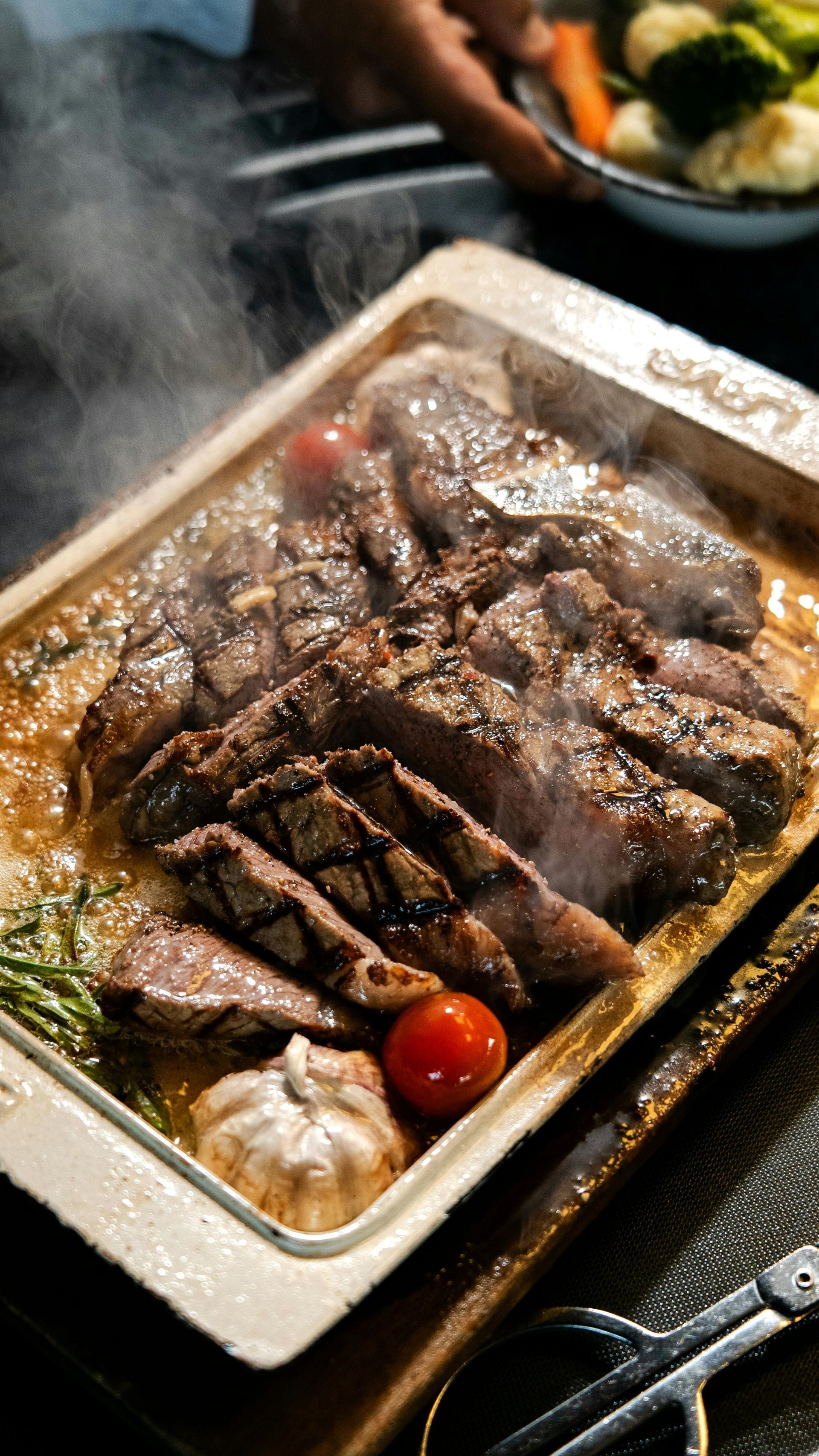 A sizzling, sliced steak served on a hot rectangular platter with a cherry tomato, garlic, and fresh herbs.