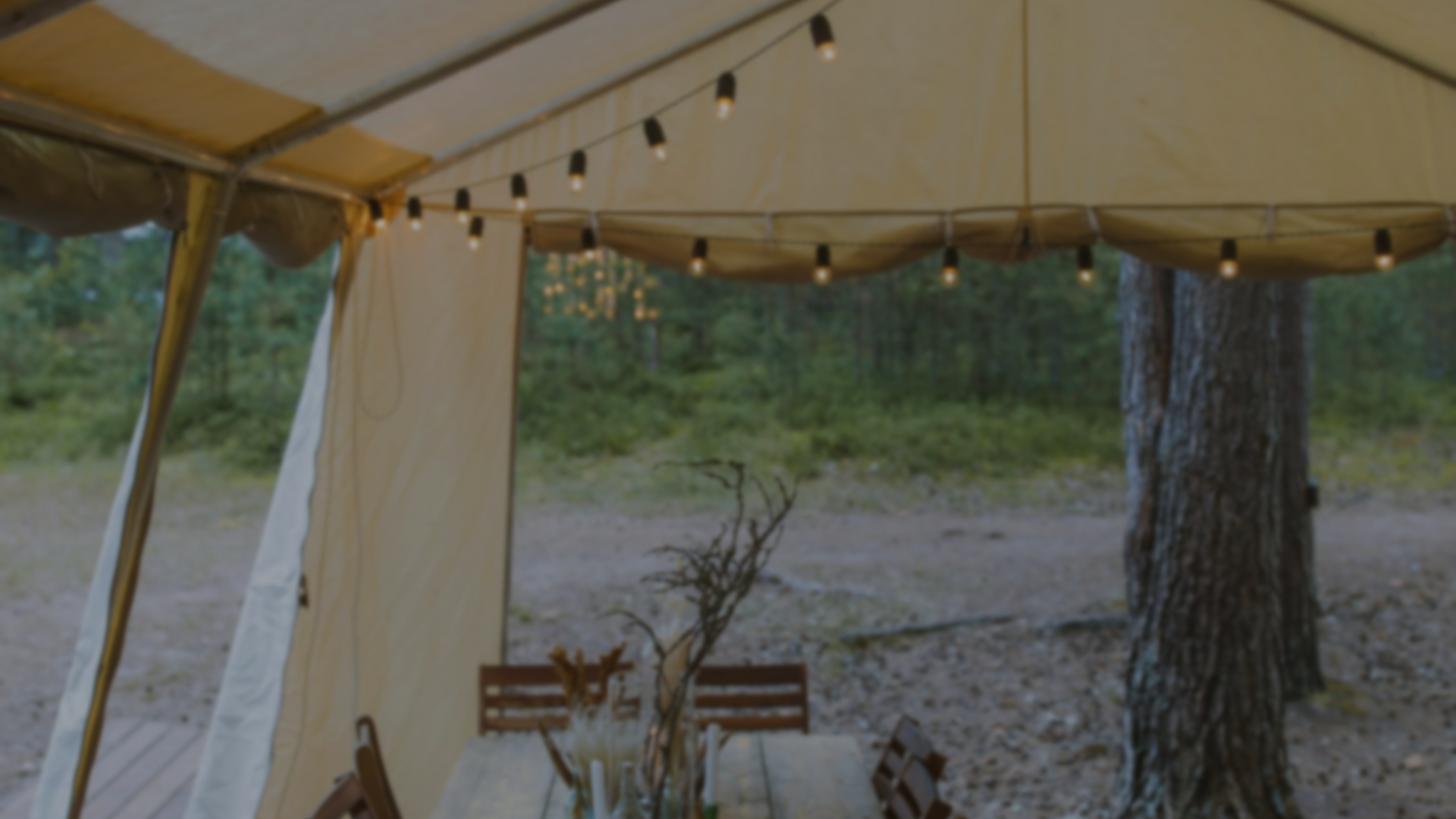 A cozy, lit outdoor tent with string lights, a table, and chairs, set against a wooded background.