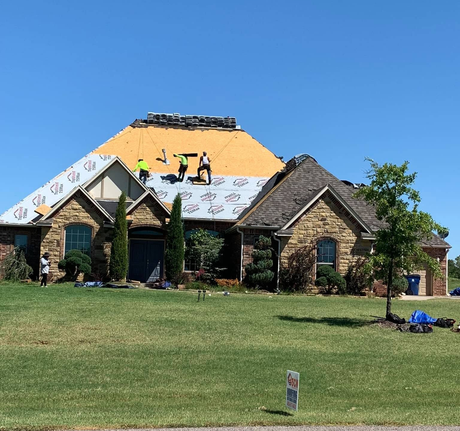 Stone house under construction with workers on the roof, bright orange sheathing, and grassy front yard.