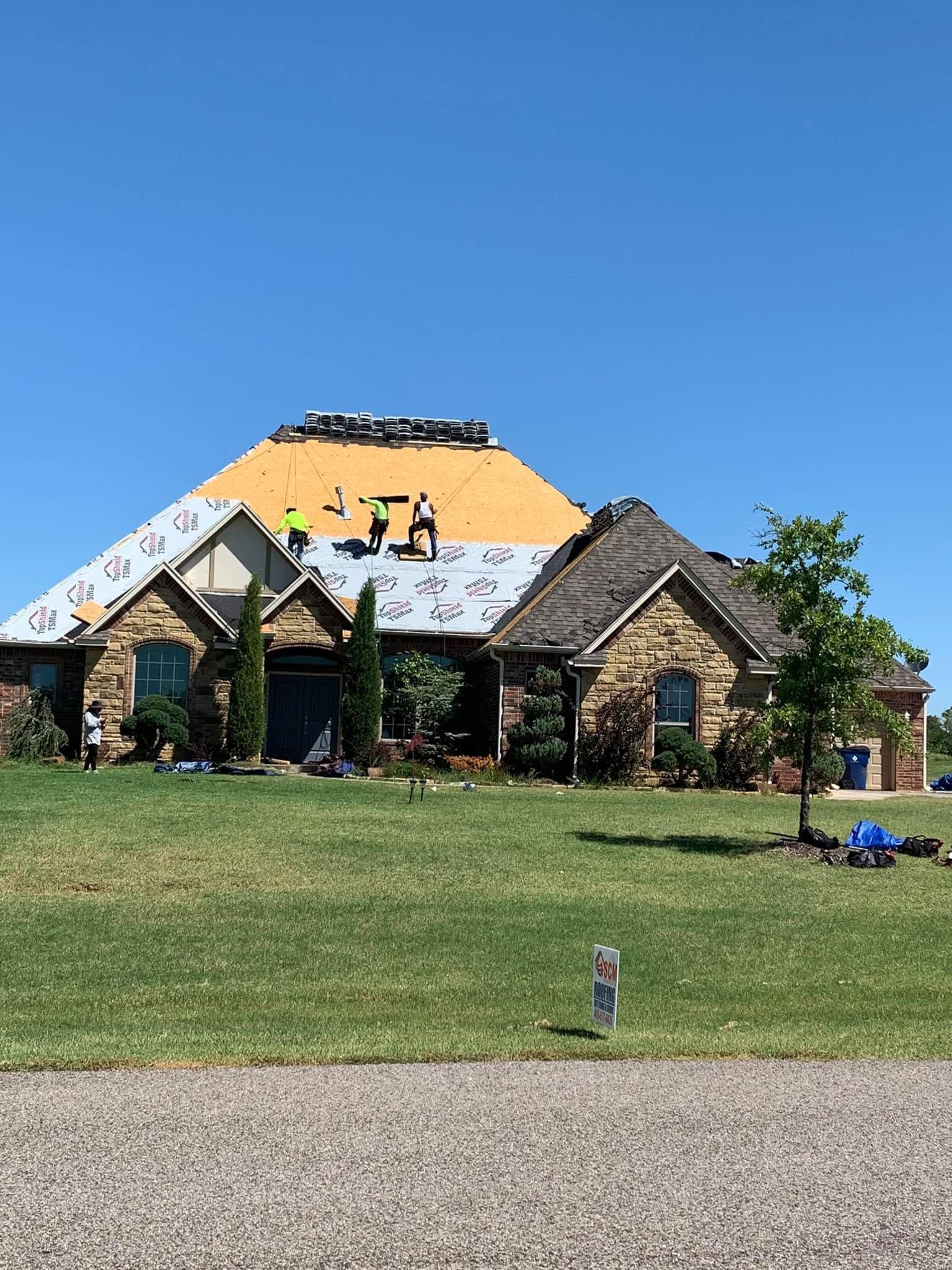 Partially roofed stone house under construction with yellow sheathing, grassy yard, and blue sky