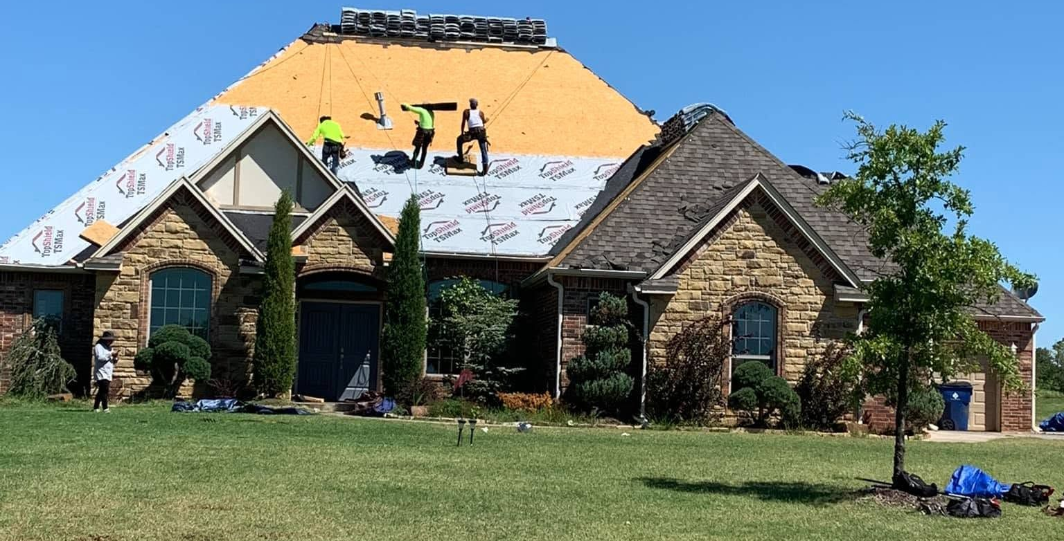 House roof under construction with workers, yellow underlayment, and shingles on a suburban home