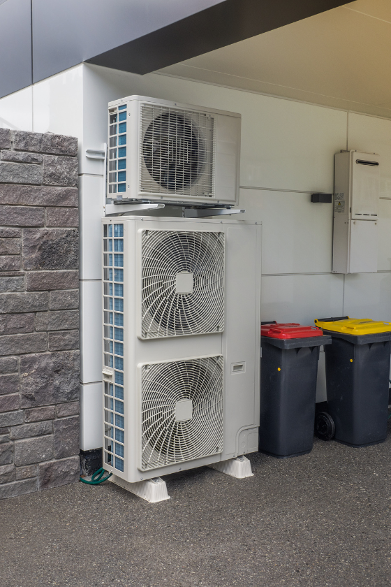 A large air conditioner is sitting outside of a building next to trash cans.