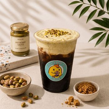 Pistachio sauce jar, iced dessert drink, and pistachios on a tabletop with leafy decor