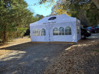 White outdoor event tent with arched windows on a wooded gravel lot, parked cars nearby