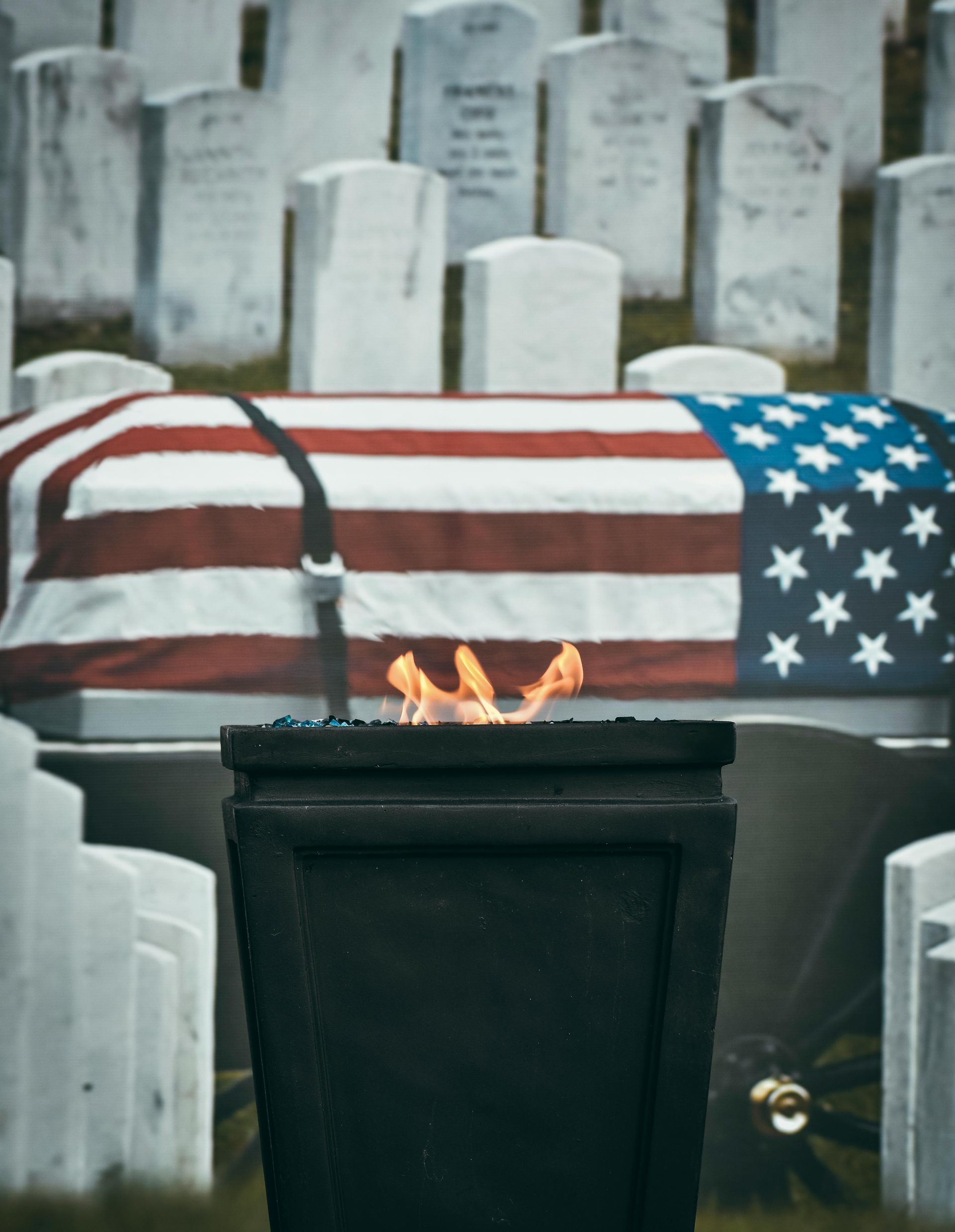 American flag-draped casket with a flame in front, surrounded by gravestones