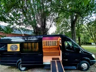 Black van parked on grass with side door open, revealing a lit wood-paneled interior and ramp under trees