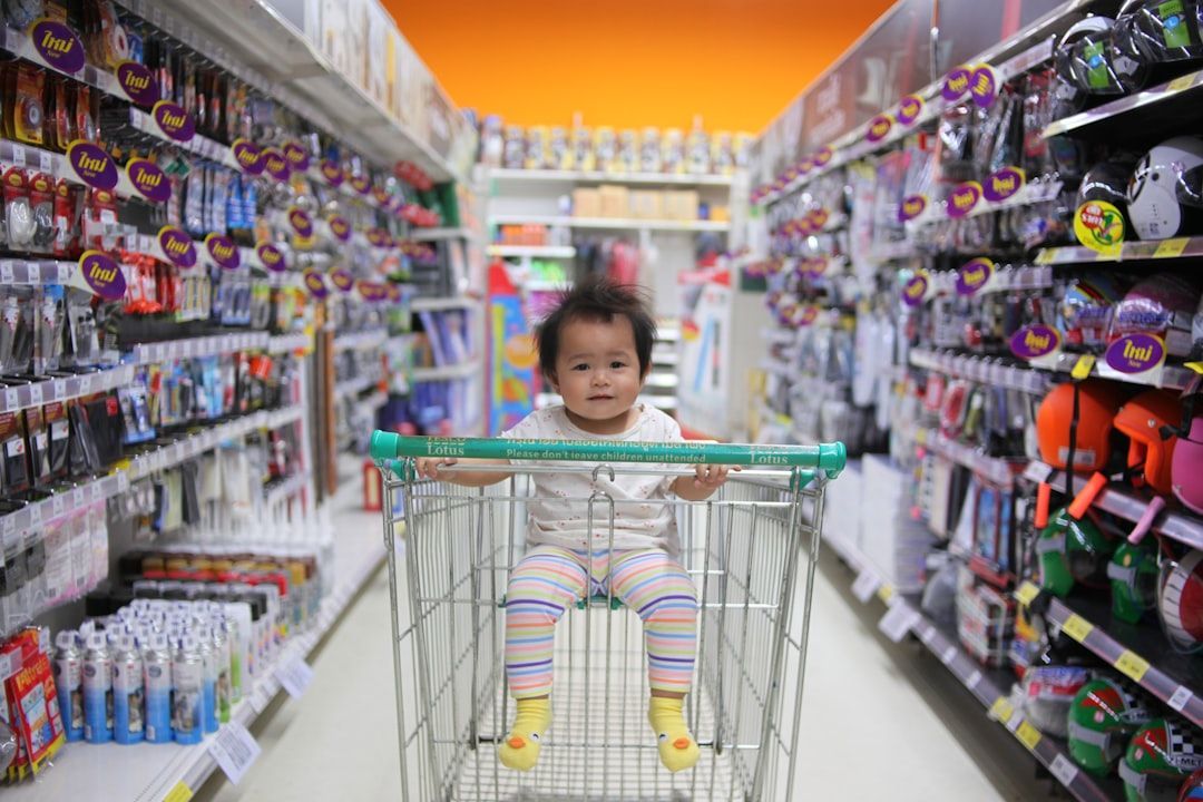 A baby sits in a shopping cart in a store aisle, looking forward with a neutral expression.