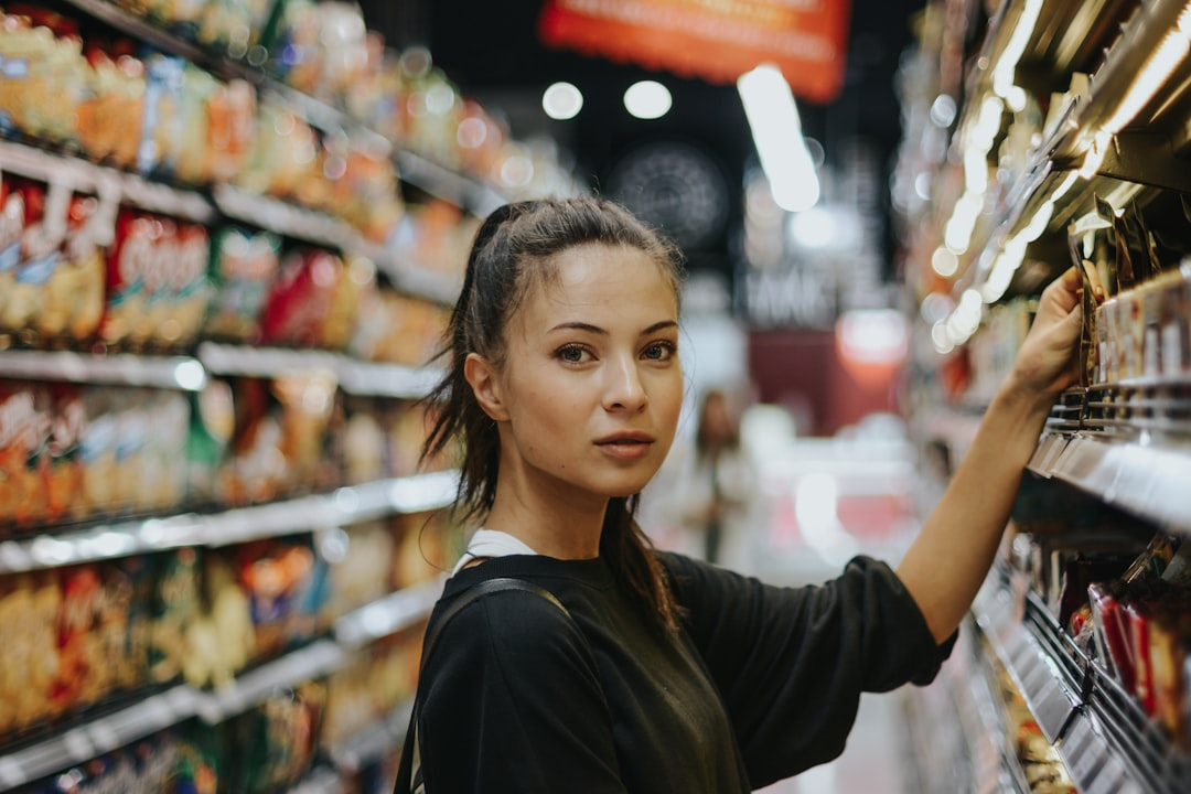 Woman shopping in a grocery store, reaching for items on a shelf. She has dark hair, a neutral expression.