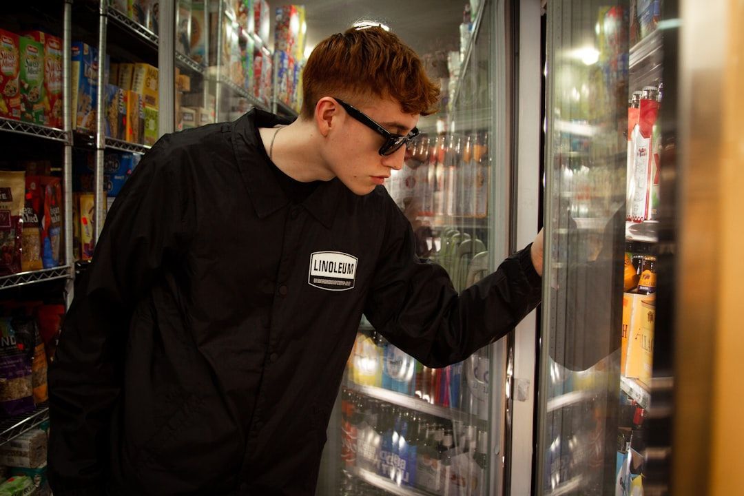 A young man with red hair and sunglasses peers into a refrigerated display case, searching for a product.