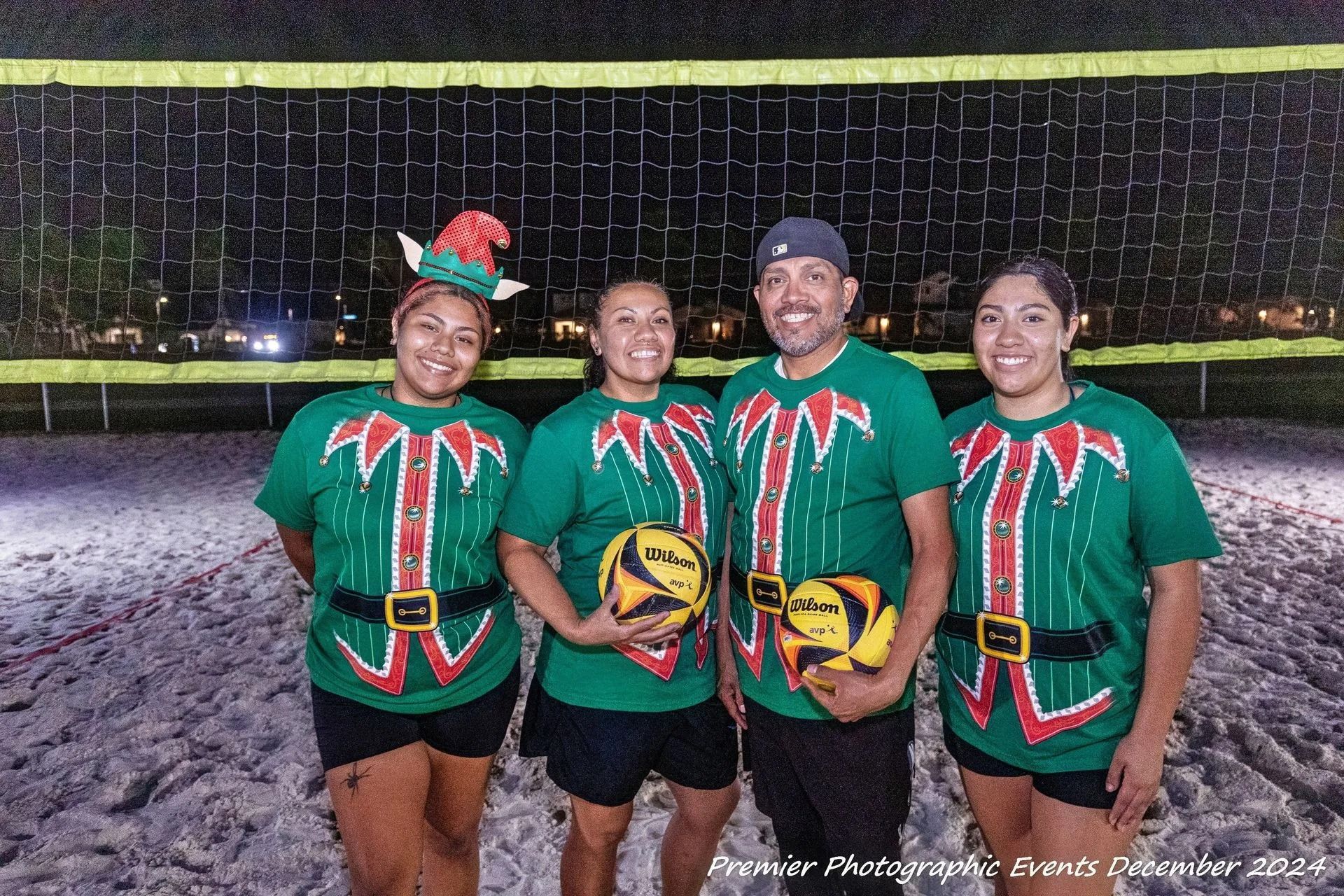 Four people in elf costumes pose on a beach volleyball court at night.