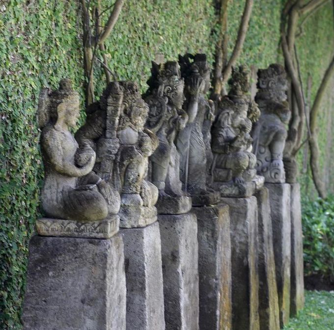 Row of weathered stone statues on pedestals beside a moss-covered wall in a lush garden