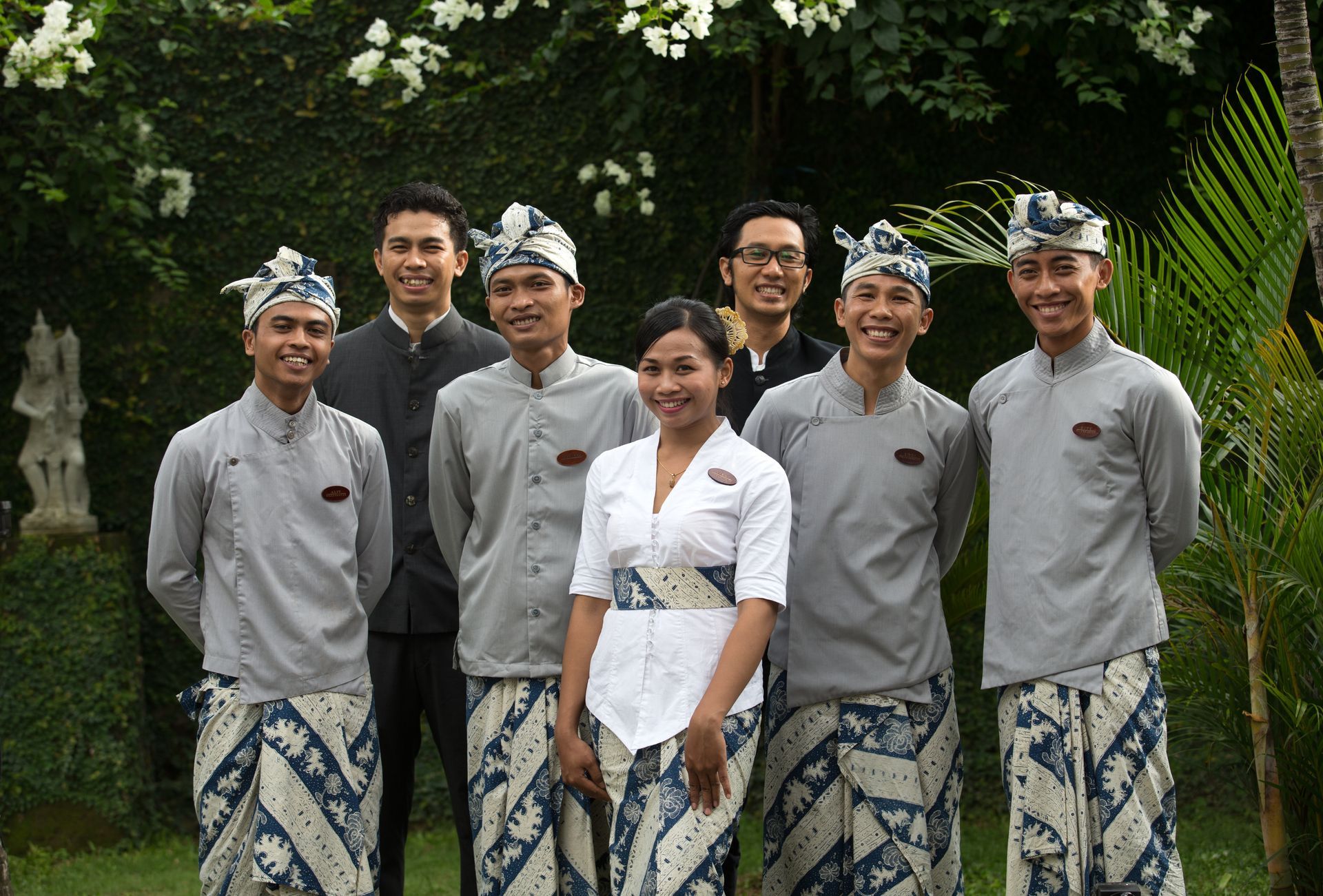 Seven people in matching traditional attire standing outdoors in front of lush greenery